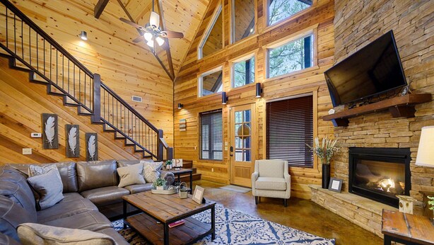 A spacious living room with a wooden interior, a fireplace, a large TV mounted on the wall, and a staircase leading to upper floors. - Luxury Broken Bow Cabin w/ Game Room & Hot Tub | Sleeps 16 (HOCHATOWN)