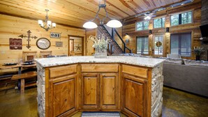 A wooden cabin interior with a stone island counter, wooden cabinets, a dining area, and a staircase leading to a loft. - Luxury Broken Bow Cabin w/ Game Room & Hot Tub | Sleeps 16 (HOCHATOWN)