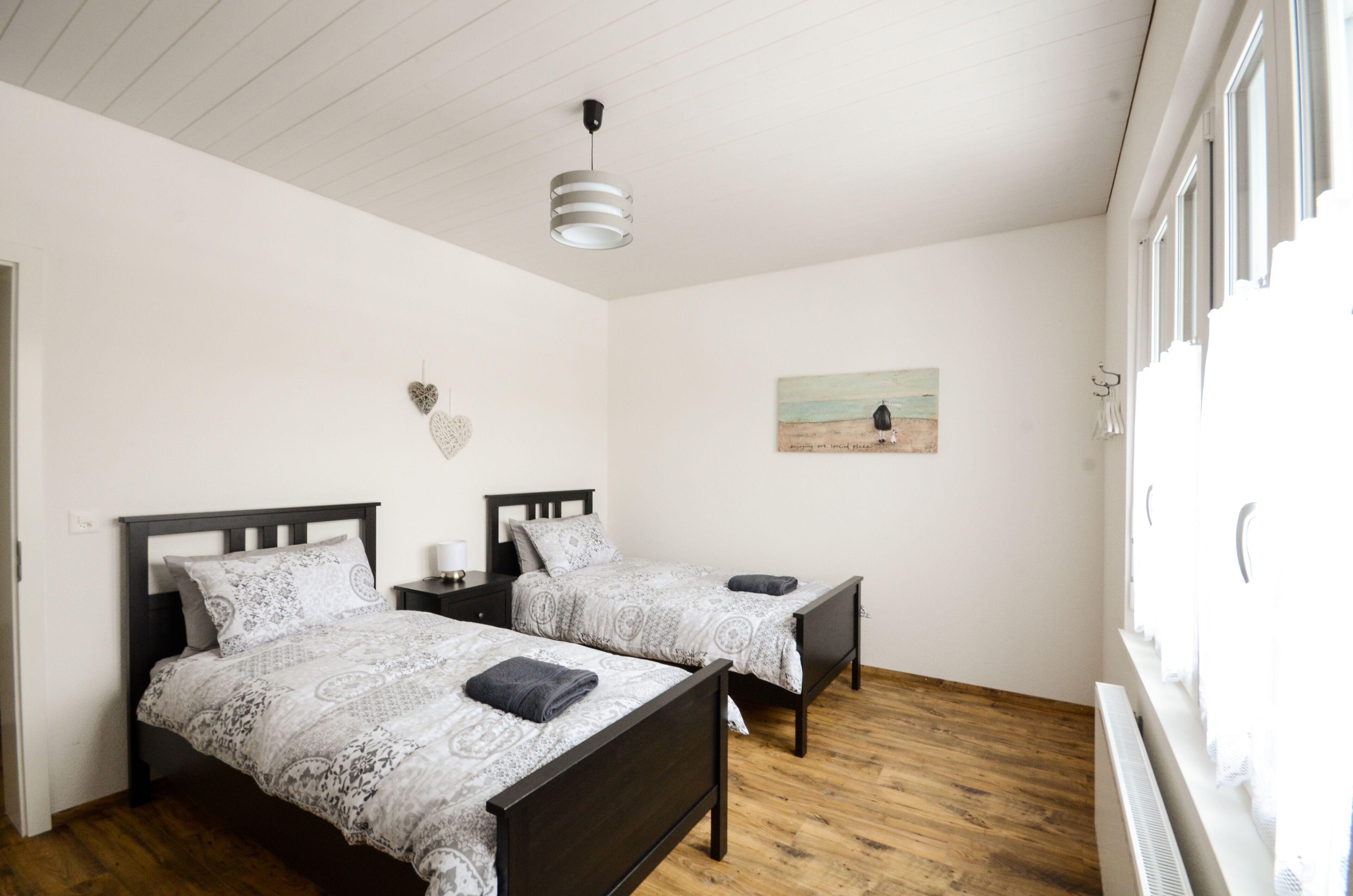 A bedroom with two beds, a ceiling light, a painting, and a window with curtains.
