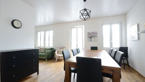 A spacious dining area with a wooden table and chairs, a black dresser, a green floral armchair, and a clock on the wall.