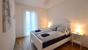 A bedroom with a white bed, a nightstand with a lamp, a window with sheer curtains, and a wooden floor.