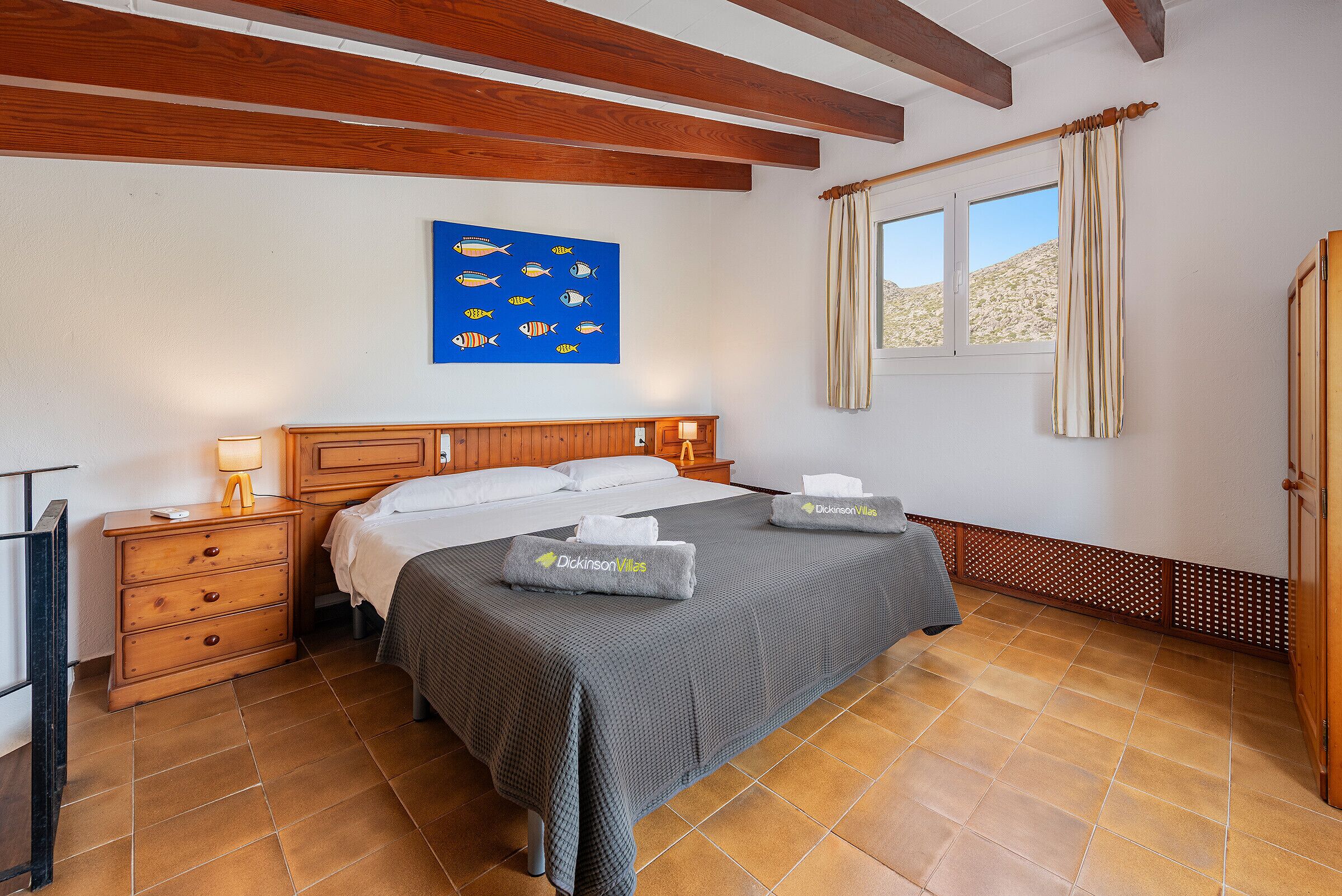A hotel room with a bed, wooden headboard, a nightstand with a lamp, a wall-mounted artwork of fish, and a window with curtains.