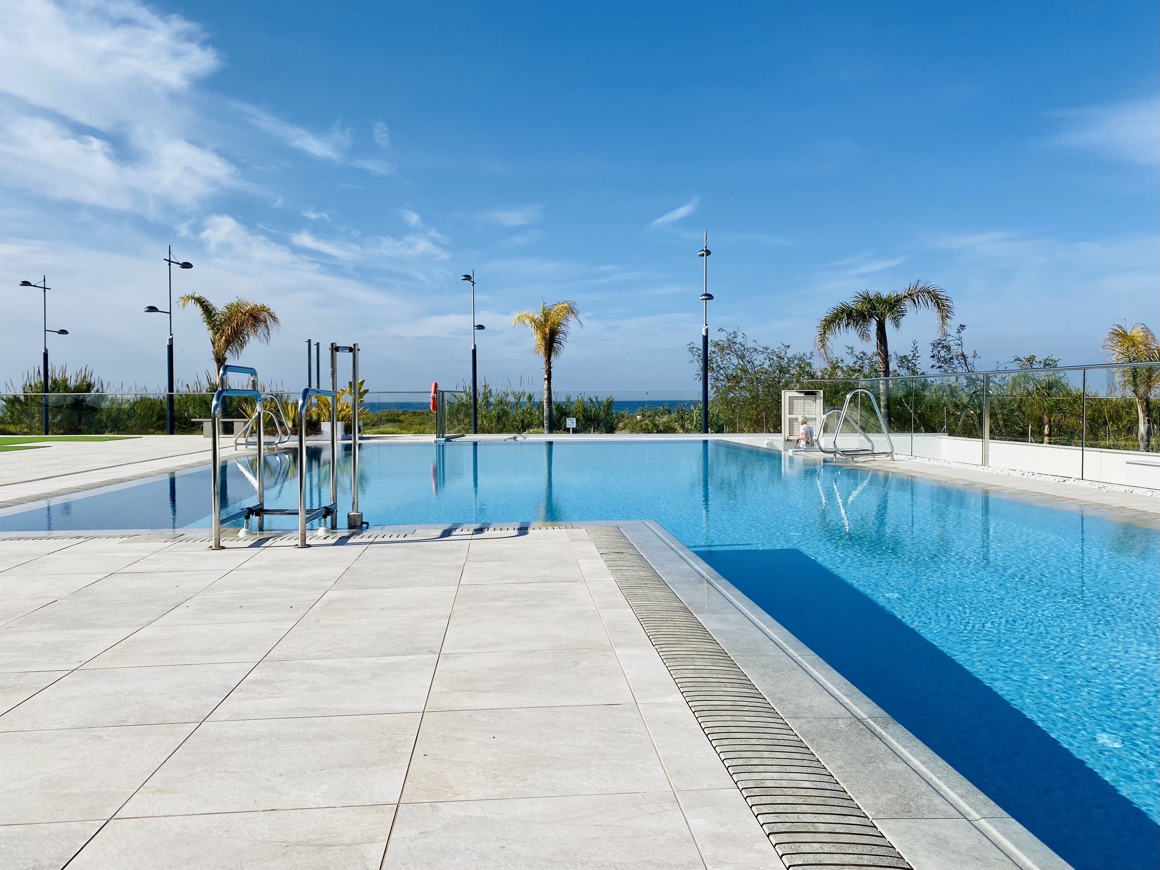 Une piscine d’un bleu éclatant, bordée d’une terrasse carrelée, entourée de palmiers et d’éclairages de rue, sous un ciel dégagé.