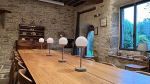 Une longue table en bois, des lampes rondes, des murs en pierre et une grande fenêtre donnant sur un paysage verdoyant.