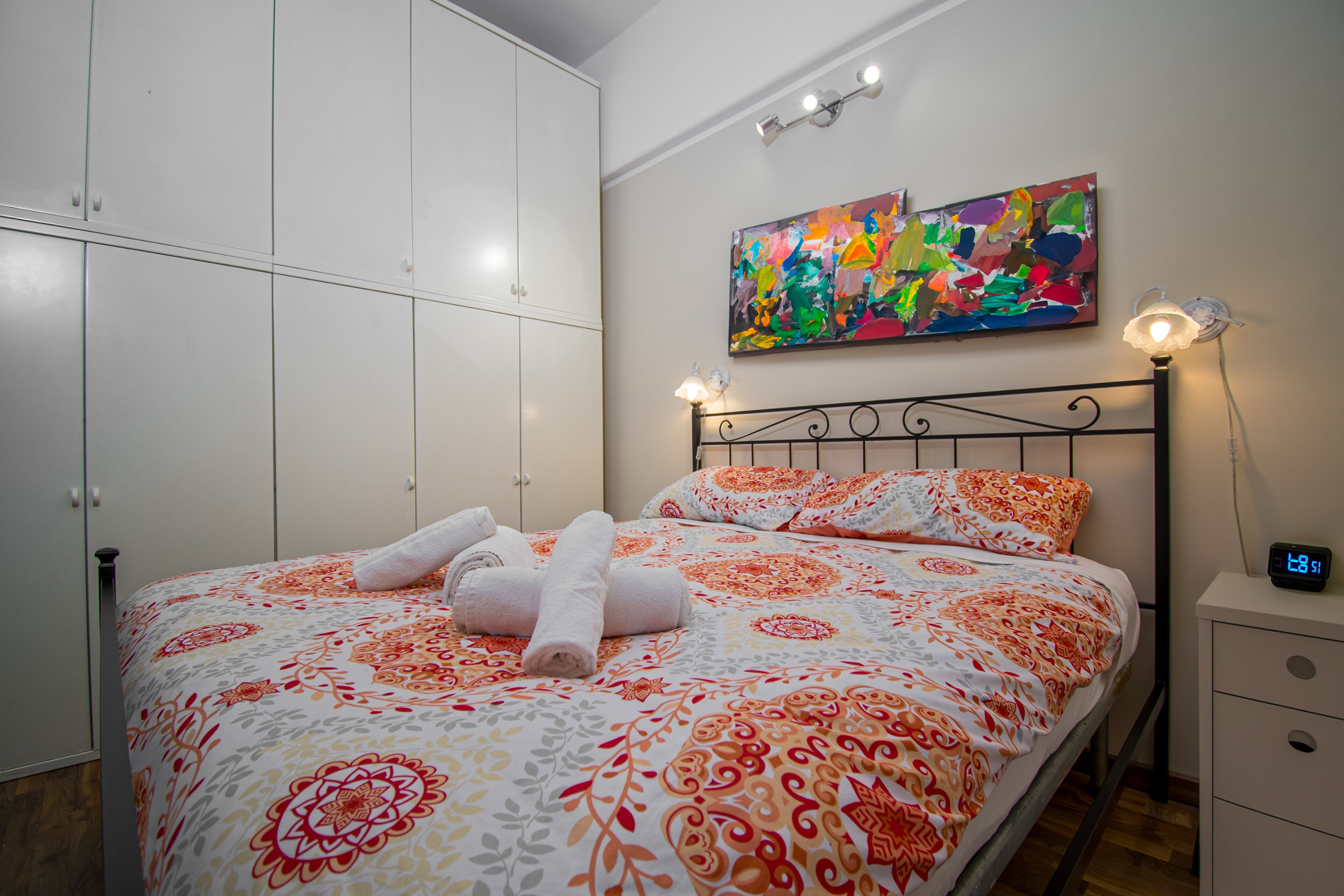 Una cama bien tendida con una colcha estampada, un cuadro en la pared y una mesita de noche con un reloj.