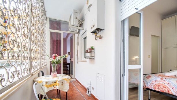 En smal balkon med et bord og en stol, en skydedør til et soveværelse og et dekorativt metalhegn.