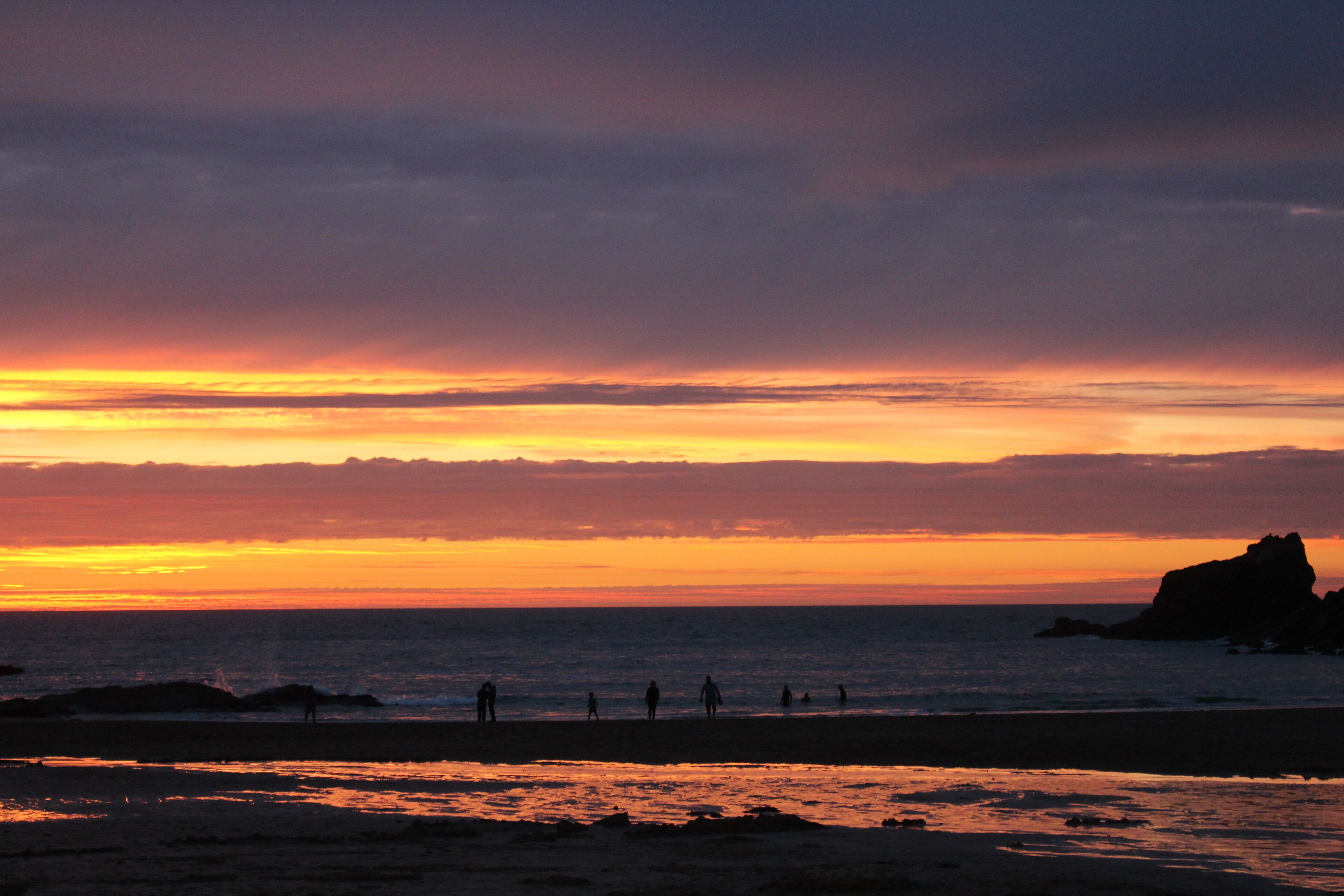 A beach at sunset with silhouettes of people and a large rock formation in the distance.