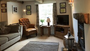 A cozy living room with a brown leather armchair, a grey sofa, a flat-screen TV, a fireplace, and framed pictures on the wall. - A Delightful 19th Century Beamed Cottage In Trevone Bay, 3 Mins From The Beach (Padstow)
