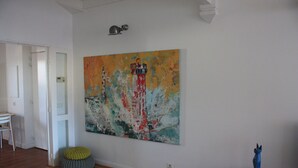 Une pièce avec un grand tableau abstrait au mur, un plancher en bois, un petit pouf rond et une sculpture bleue posée sur une table en bois.