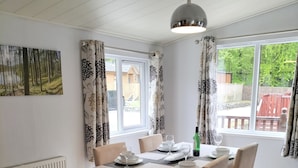 Uma área de refeições com uma mesa posta para quatro pessoas, uma luminária pendente e vista para o exterior através de grandes janelas.