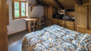 Une chambre avec un plafond en bois, un lit recouvert d’une couvre-lit à motifs floraux, un bureau avec une chaise et une fenêtre donnant sur un paysage verdoyant.