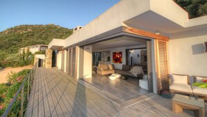 Une maison moderne dotée d’une terrasse en bois, d’un espace de vie avec des canapés et offrant une vue sur une colline verdoyante.