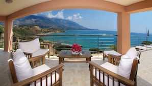 A balcony with wooden furniture and a table, overlooking a coastal landscape with mountains and the sea.