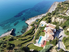 Vue aérienne d’une propriété située sur la côte, avec une piscine, une plage et des jardins aménagés en terrasses.
