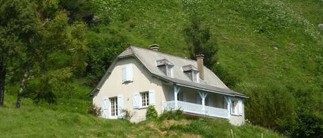 Een huis met schuine kap, witte gevel en een balkon. Het staat op een groene heuvel, omringd door bomen.