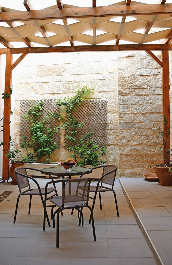 Une terrasse couverte avec une table et des chaises, entourée de plantes en pot et d’un mur de pierre.