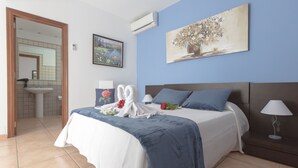 A neatly made bed with a blue and white bedspread, a white towel, and a bouquet of flowers. There is a painting on the wall above the bed, a bedside lamp, and a view of a bathroom with a sink and mirror. -  VILLA "BELLA LUCIA 1" PRIVATE HEATED POOL, WIFI, BARBECUE, SATELLITE TV  (corralejo- la oliva)