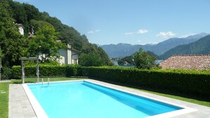 A rectangular swimming pool with a white border, surrounded by a paved area and a well-maintained hedge. In the background, there are houses with red-tiled roofs, a mountain range, and a clear blue sky. - “For a perfect and UNFORGETTABLE GIFT … (Sala Comacina)