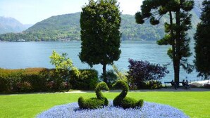 A landscaped garden with a circular flower bed, two topiary swans, and a lake with mountains in the background. - “For a perfect and UNFORGETTABLE GIFT … (Sala Comacina)