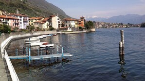 A lakeside town with buildings along the shore, a dock with boats, and mountains in the background. - “For a perfect and UNFORGETTABLE GIFT … (Sala Comacina)