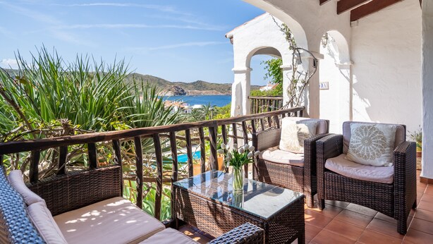 A balcony with wicker furniture, a glass table, and a view of the sea and hills. - Casa Lavanda (Fornells, Menorca)