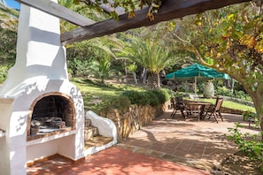 Enjoy al fresco dining under a shaded pergola with a brick oven and lush greenery.