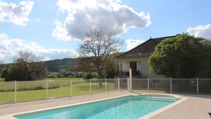 A rectangular swimming pool with a white border, surrounded by a white fence, set against a backdrop of a house and trees under a partly cloudy sky. - Holiday Home with Large Private Pool And Hill Views In The Heart Of The Dordogne (Condat-Sur-Vézère)