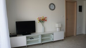 Una sala de estar moderna con un televisor de pantalla plana sobre un mueble blanco, un jarrón con flores, un reloj en la pared y una puerta cerrada.