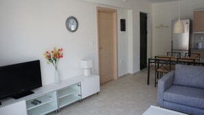 Una sala de estar moderna con un televisor de pantalla plana, una consola blanca, un jarrón con flores, un sofá gris y una zona de comedor con una mesa y sillas.
