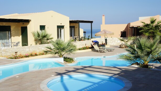 Zona de piscina con sillones de descanso y una persona trabajando en una mesa, rodeada de palmeras y un muro de piedra.