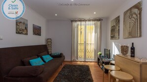 Una sala de estar con un sofá marrón, una mesita de centro y un televisor. Hay dos cuadros en la pared y una ventana con cortinas.