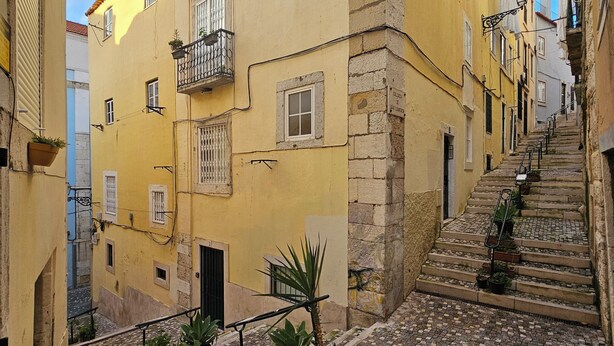 Una estrecha calle empedrada flanqueada por edificios amarillos con balcones y una escalera que sube.