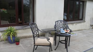 Relax in the comfort of this outdoor seating area, featuring two chairs with cushioned seats and tables, surrounded by lush greenery and a beautiful building with large windows.