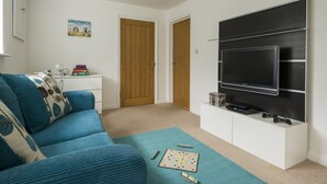 Ein Wohnzimmer mit blauem Sofa, einem Couchtisch mit einem Gesellschaftsspiel, einem flachbildfernseher an der Wand und einer Tür zu einem anderen Raum.
