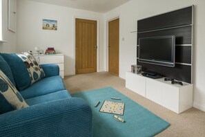 A living room with a blue sofa, a coffee table with a board game, a flat-screen TV mounted on the wall, and a door leading to another room. - Rock House - sleeps 8 guests  in 4 bedrooms (Bude)