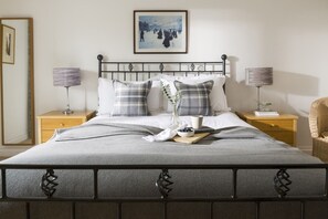 A neatly made bed with a grey blanket, two bedside tables with lamps, a framed picture on the wall, and a wicker chair. - Rock House - sleeps 8 guests  in 4 bedrooms (Bude)