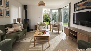 Ein Wohnzimmer mit Sofa, Couchtisch und Fernseher. Ein Kamin, ein Fenster mit Vorhängen und ein Bücherregal sind ebenfalls vorhanden.