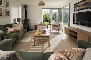 A living room with a sofa, coffee table, and TV. There is a fireplace, a window with curtains, and a bookshelf. - Rock House - sleeps 8 guests  in 4 bedrooms (Bude)