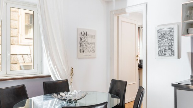 Un coin repas avec une table en verre, des chaises noires et un mur blanc orné d’œuvres d’art encadrées.