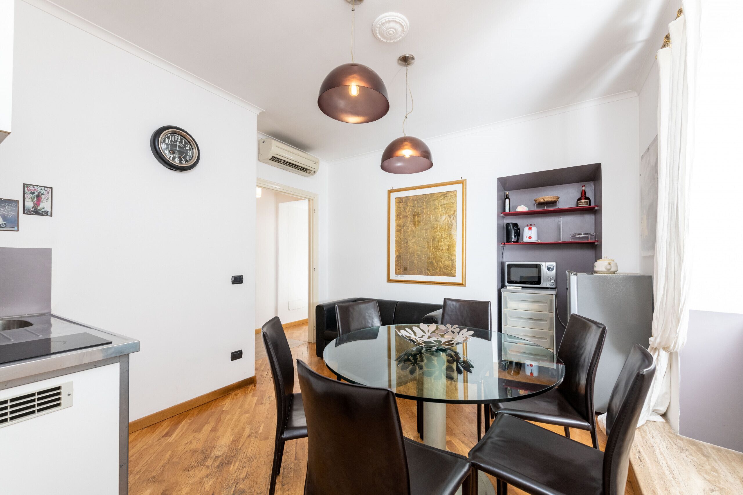A modern kitchen with a round glass table, black chairs, pendant lights, and a microwave.