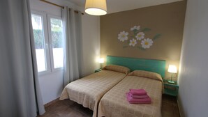 Un llit doble amb un edredó beig estampat, dues taules de nit amb làmpades, una finestra amb cortines i una paret amb un mural floral.