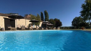Piscina con sillones de descanso y sombrillas, rodeada de edificios de piedra y árboles.