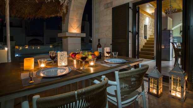 Un espacio exterior para comer con una mesa dispuesta para dos, rodeado de sillas de mimbre e iluminado con velas y faroles.