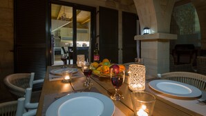 Una mesa de comedor preparada para cuatro personas, con platos, cubiertos y vasos, rodeada de sillas de mimbre. Hay un frutero y velas sobre la mesa.