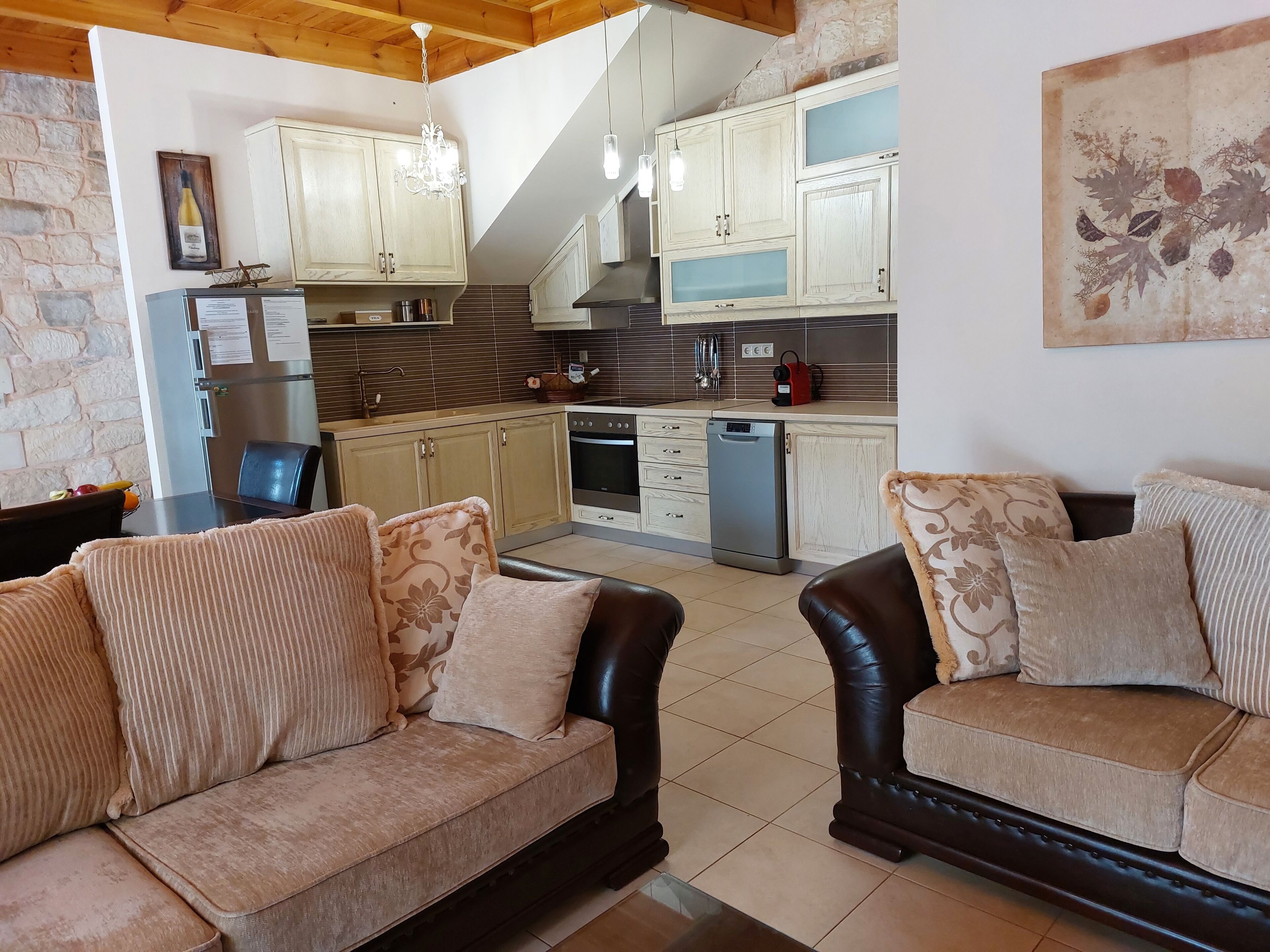 A living room with two sofas, a dining table with fruits, a kitchen with wooden cabinets, and a stone wall.