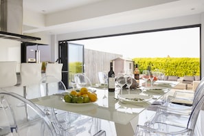 A modern dining area with a glass table, clear chairs, and a view of a lush garden. - STUNNING CONTEMPORARY COASTAL HOLIDAY HOME WITH HOT TUB, SEA VIEWS AND PARKING (Newquay)