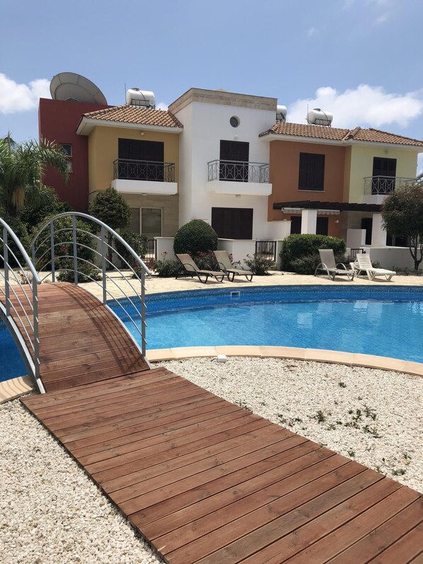 A swimming pool with a wooden deck, lounge chairs, and a building with balconies. - Beautiful Holiday Villa With Pool In Tombs Of The Kings Area . Sleeps 4 . (kato Paphos)
