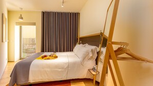 A modern hotel room with a large bed, a wooden hanger, a bedside table, and a view of the outdoors through a window.