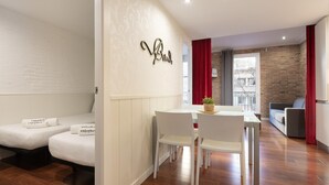 A modern hotel room with a white bed, a dining area with a table and chairs, and a brick wall.