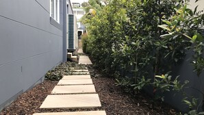 Um caminho estreito ladeado por um muro cinzento e vegetação densa, conduzindo a um edifício com aparelhos de ar condicionado.